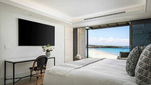 Quarto com cama grande, escrivaninha e cadeira. Há uma TV na parede e uma janela com vista para a praia.