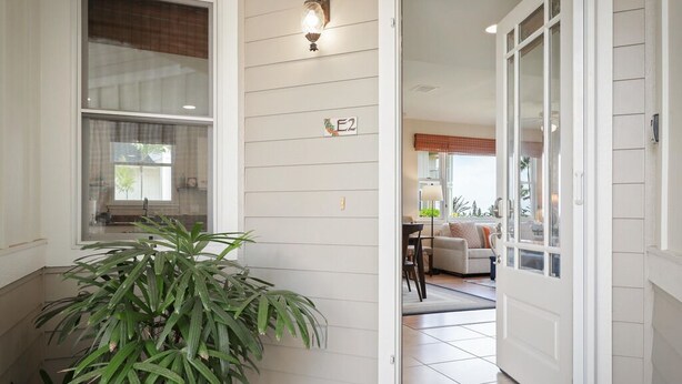 Une entrée d’immeuble d’habitation agrémentée d’un pot de fleurs, une porte avec une plaque de numéro et un aperçu d’un salon avec un canapé et une lampe.