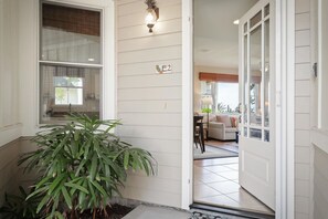 A residential entrance with a potted plant, a door with a number plate, and a glimpse into a living room with a sofa and a lamp.