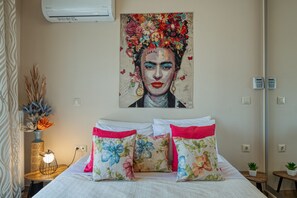 Un dormitorio con cama, almohadillas estampadas, un cuadro, una mesita de noche con lámpara y un aire acondicionado de pared.