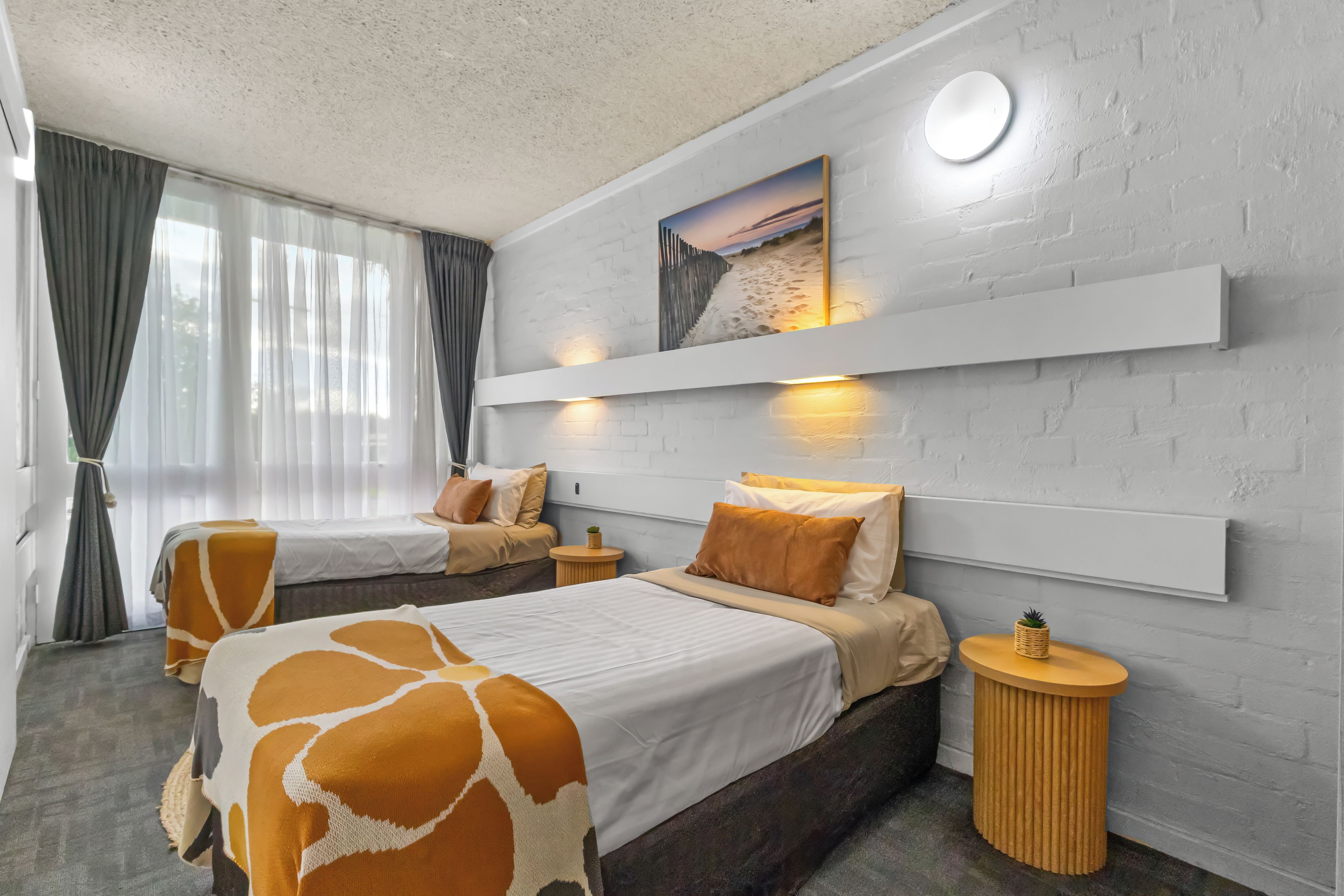 A hotel room with two beds, a large window with curtains, a wall-mounted shelf with a picture, and a round light fixture.