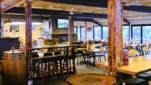 A restaurant with wooden tables and chairs, a bar area, and a television screen displaying a program. - Mallacoota Hotel Motel (Mallacoota)