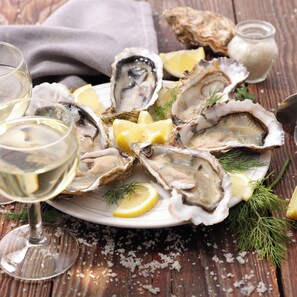 A plate of oysters with lemon wedges and dill, accompanied by a glass of white wine on a wooden table. - Mallacoota Hotel Motel (Mallacoota)