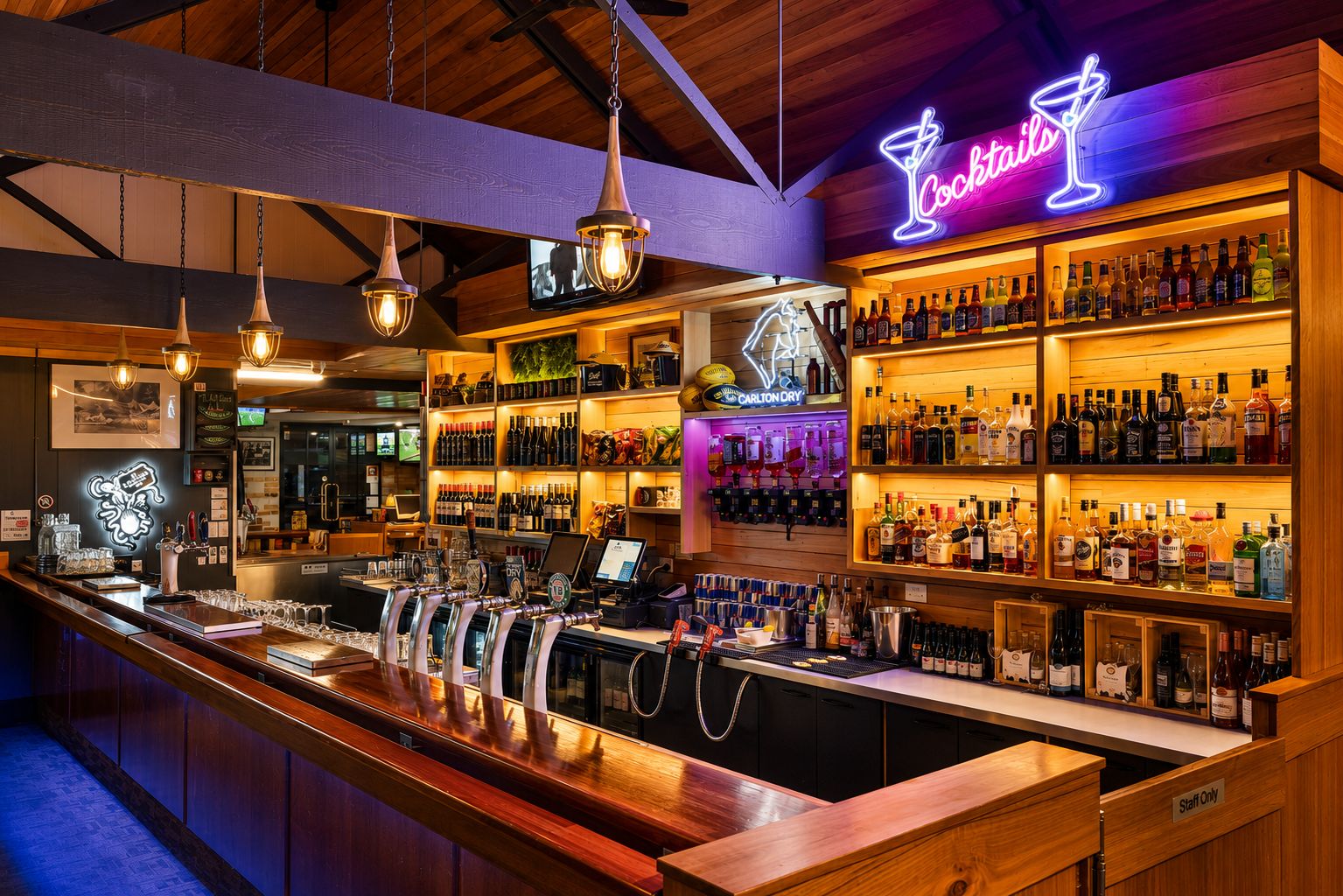 Zona de bar con un letrero de cócteles de neón, varias estanterías llenas de botellas de licor y una barra con grifos y vasos.