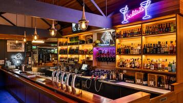 Zona de bar con un letrero de cócteles de neón, varias estanterías llenas de botellas de licor y una barra con grifos y vasos.