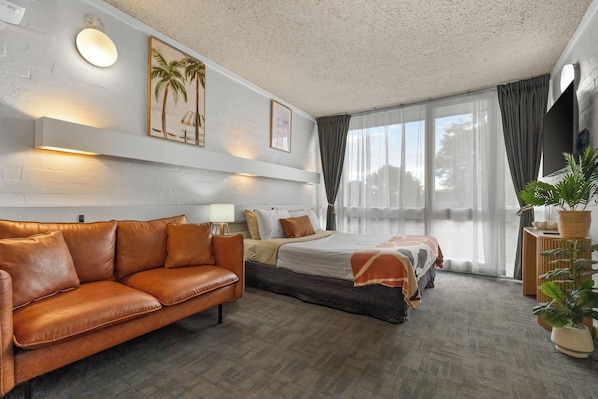 Habitación de hotel con sofá, cama, mesita de noche, lámpara, planta en maceta y ventana con cortinas.