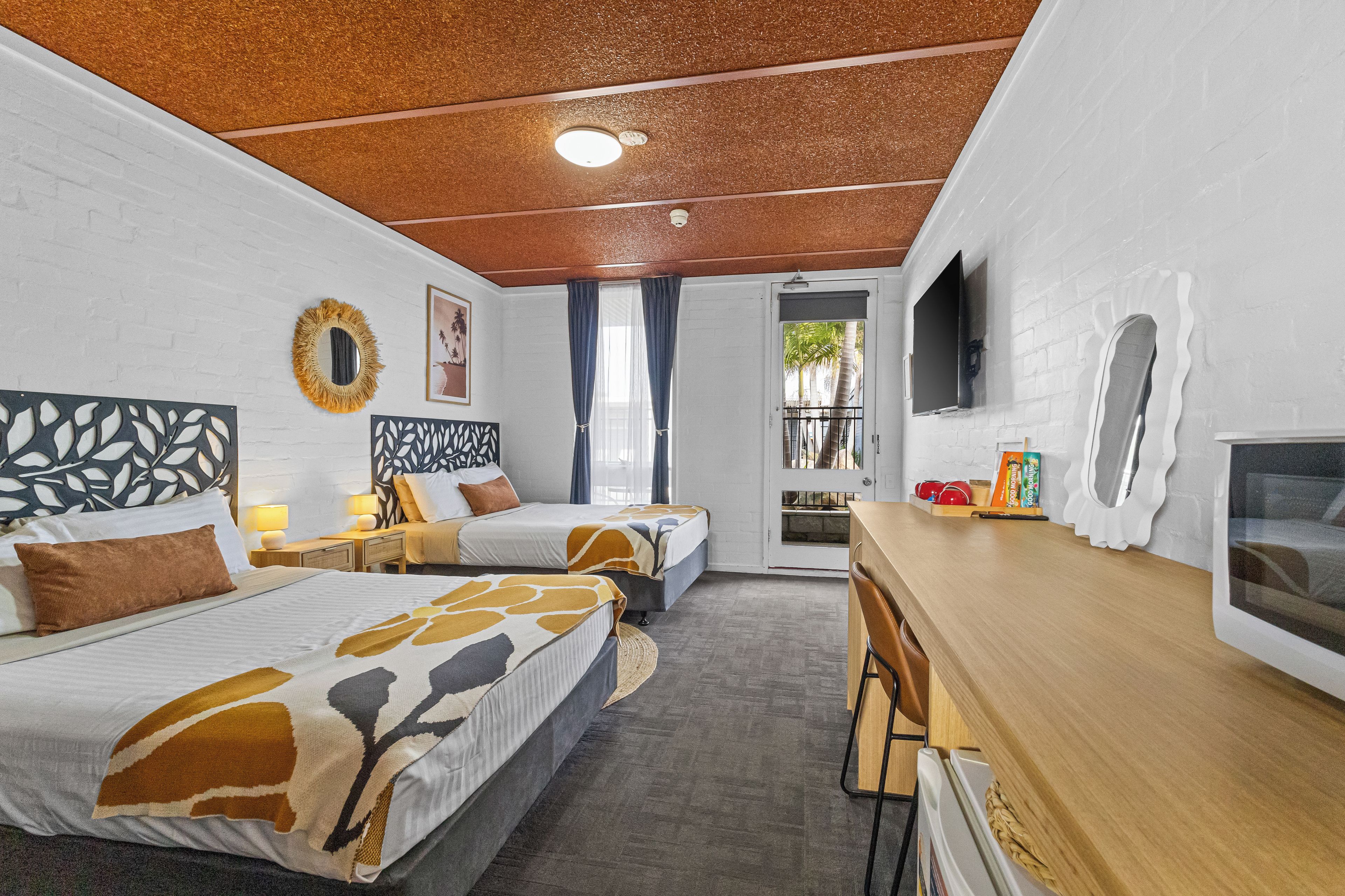 A hotel room with two beds, a desk, a TV, and a balcony with a view of palm trees.