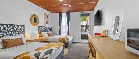 A hotel room with two beds, a desk, a TV, and a balcony with a view of palm trees.