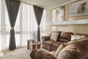 A living room with a brown leather sofa, a small round table, and a large window with curtains. - Mallacoota Hotel Motel (Mallacoota)