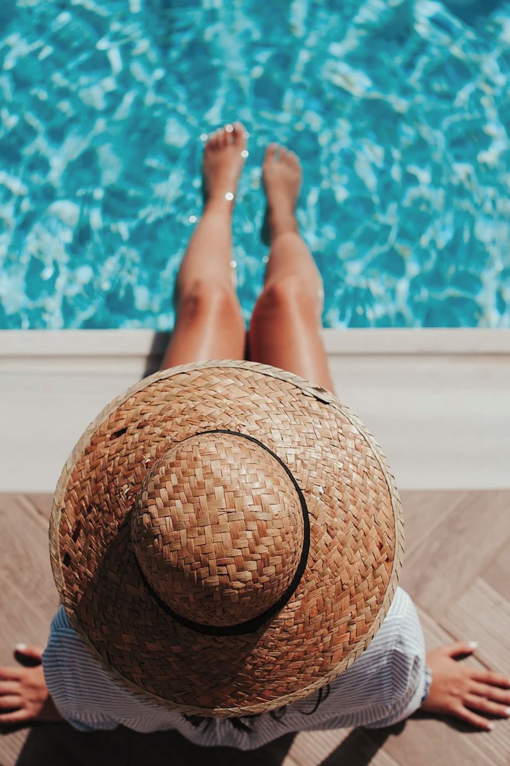 Una persona sdraiata su una poltrona a bordo piscina, con un cappello di paglia, che guarda la piscina.