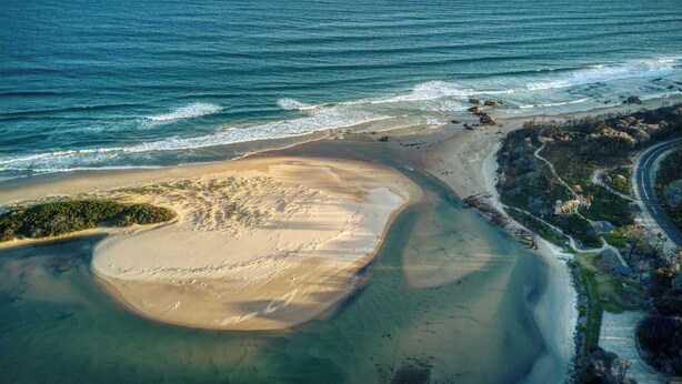 An aerial view of a coastal area with a sandy beach, clear blue waters, and a road curving along the coast. - Mallacoota Hotel Motel (Mallacoota)