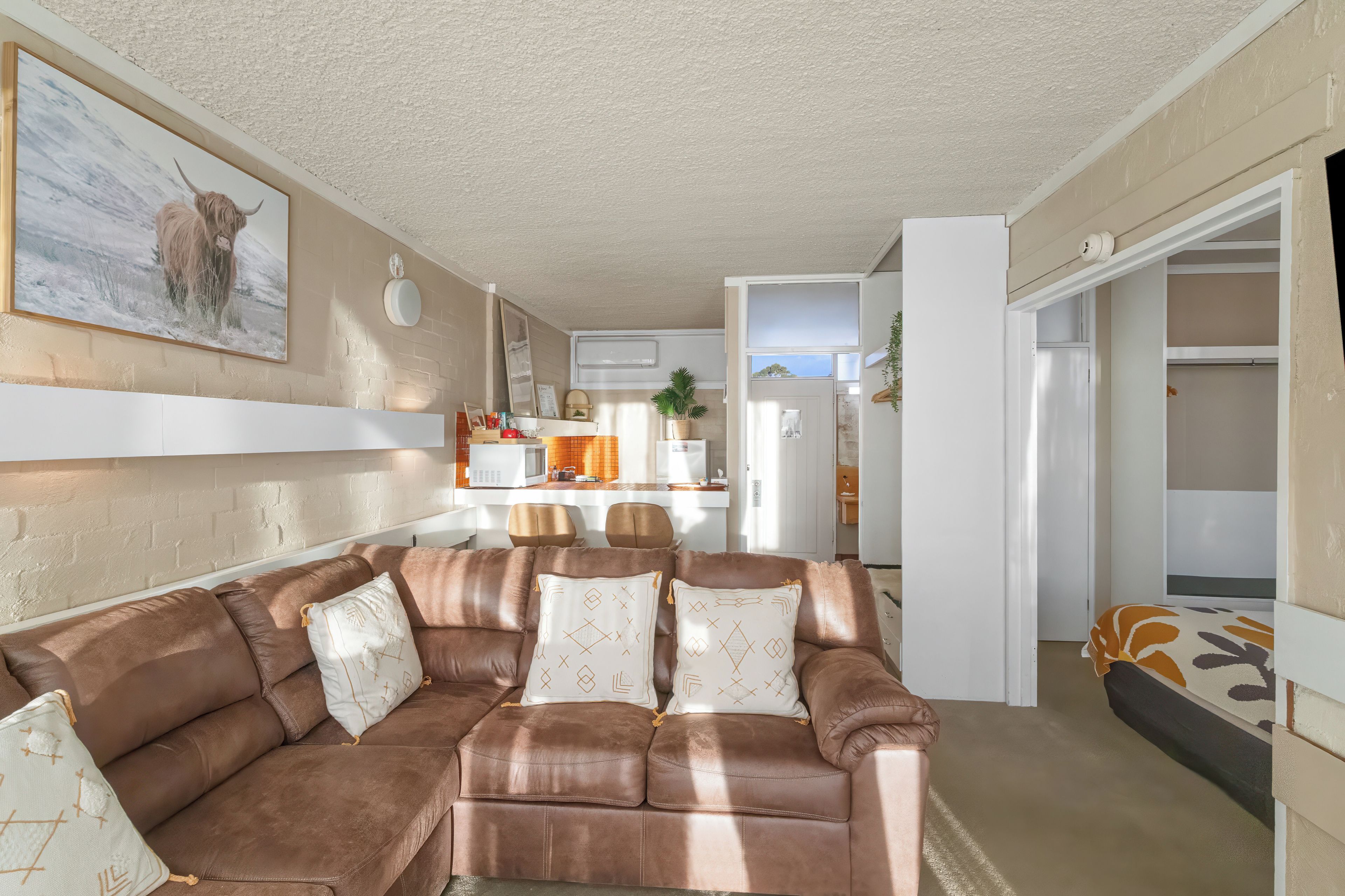 Sala de estar com sofá de couro marrom, cozinha ao fundo e área de jantar.