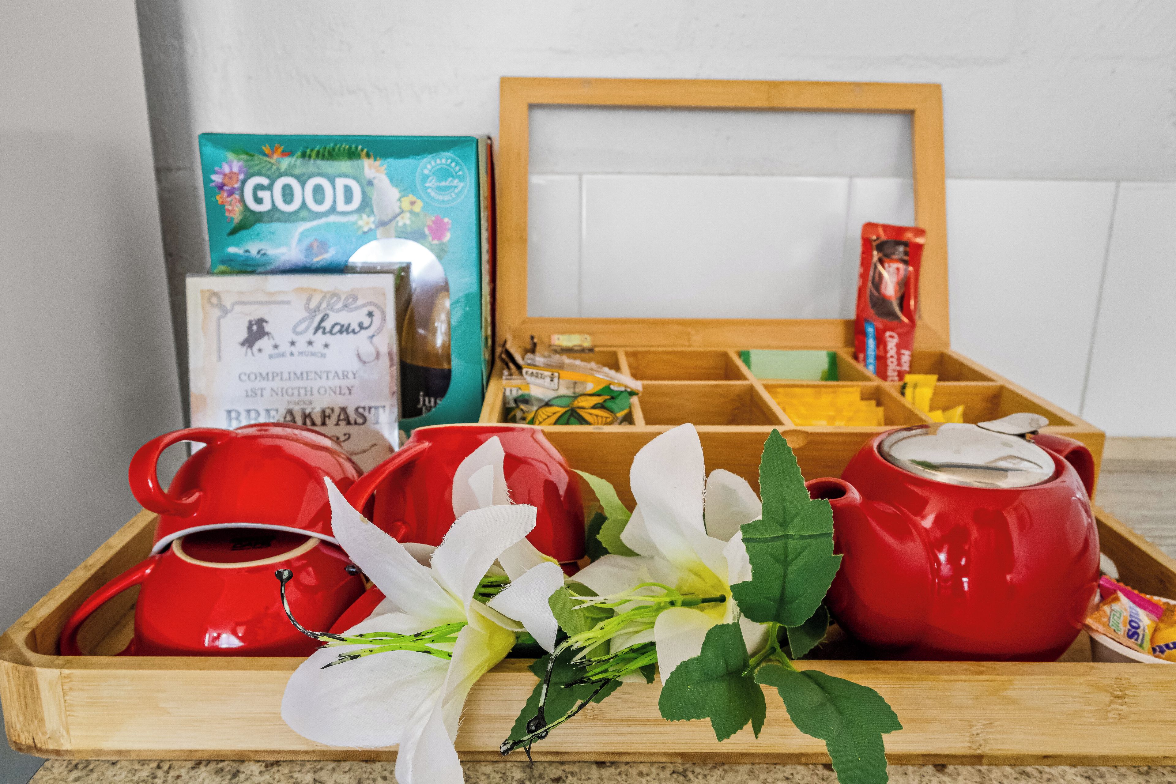 A breakfast tray with red teapots, a teapot, a cup, a box of 'GOOD' tea, a 'We Shaw' branded item, and a bouquet of flowers.