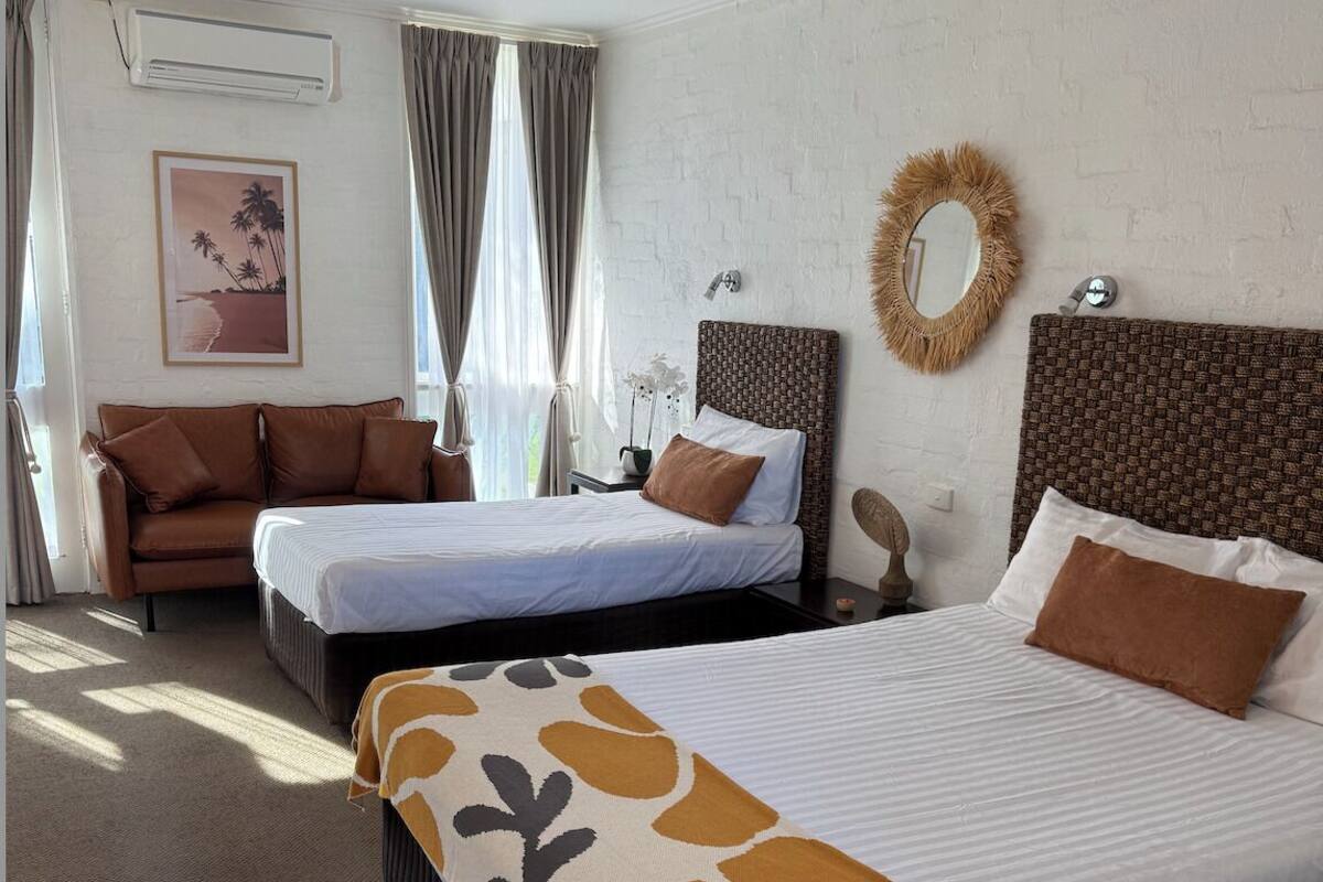 A hotel room with two beds, a sofa, a mirror, and a wall-mounted air conditioner.