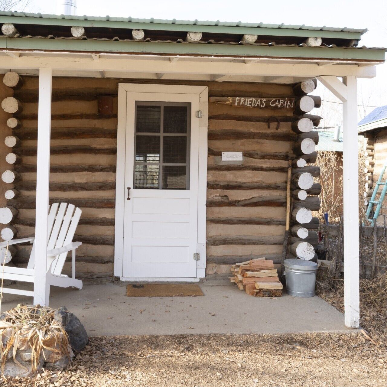 Una cabaña de troncos con una puerta blanca, un letrero que dice 'FRIEDAS CABIN' y una silla blanca afuera.