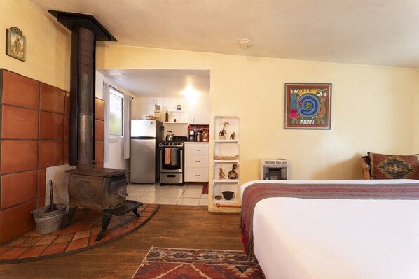 A room with a bed, a wooden stove, a kitchen with a refrigerator and oven, and a decorative wall piece.