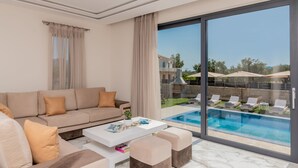 Un soggiorno moderno con un divano beige, un tavolino da caffè bianco e un'ampia porta scorrevole in vetro che dà su una zona piscina.
