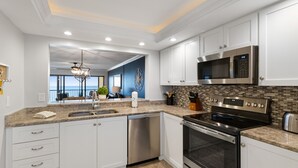 A modern kitchen with white cabinets, stainless steel appliances, and a view to the outside. - 🌅 Ocean Sands II #606 — Gulf Front Elegance with Sunset Views (Madeira Beach)