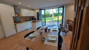 Una cocina moderna con armarios blancos, un comedor con mesa de madera dispuesta para cuatro personas, y vista a un jardín a través de grandes puertas corredizas de vidrio.