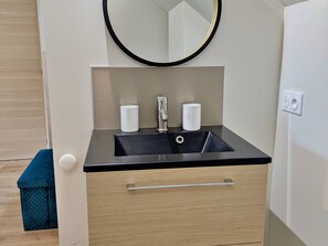 Un baño moderno con un lavamanos negro, dos vasos blancos, un espejo y un mueble de madera con un cajón.