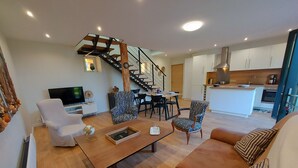 Una sala de estar moderna con una mesa de centro de madera, sillones blancos, sillas estampadas y una escalera que lleva a una zona de cocina.