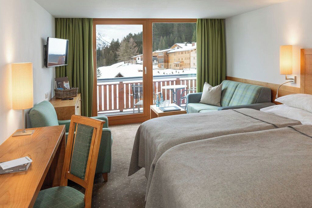 Habitación de hotel con dos camas, un escritorio, una silla, un televisor y un balcón con vistas a una montaña nevada y edificios.