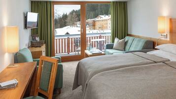 Habitación de hotel con dos camas, un escritorio, una silla, un televisor y un balcón con vistas a una montaña nevada y edificios.