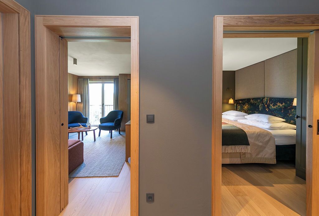 A hotel room with a bed, a desk, and a chair. There is a view of the hallway and another room.