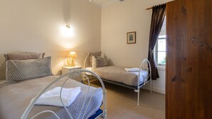A bedroom with two beds, a bedside table with a lamp, a window with curtains, and a wooden door. - Bluebush Estate - The Cottage (Rothbury)