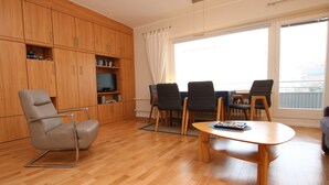 Uma sala de estar moderna com piso de madeira, uma poltrona reclinável cinza, uma mesa de jantar com quatro cadeiras e uma TV em uma estante.