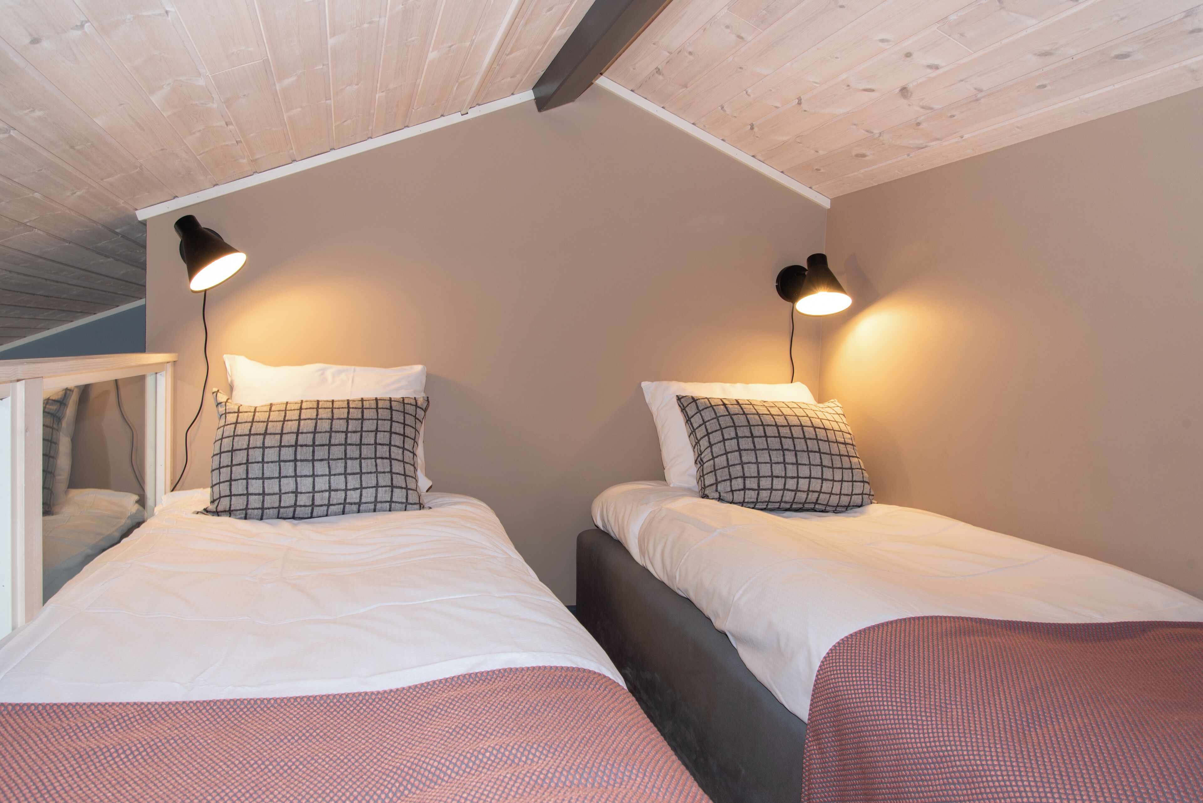 Un dormitorio con dos camas, cada una con cabecero, ropa de cama blanca y almohada estampada. Hay dos lámparas fijadas a la pared que brindan luz.