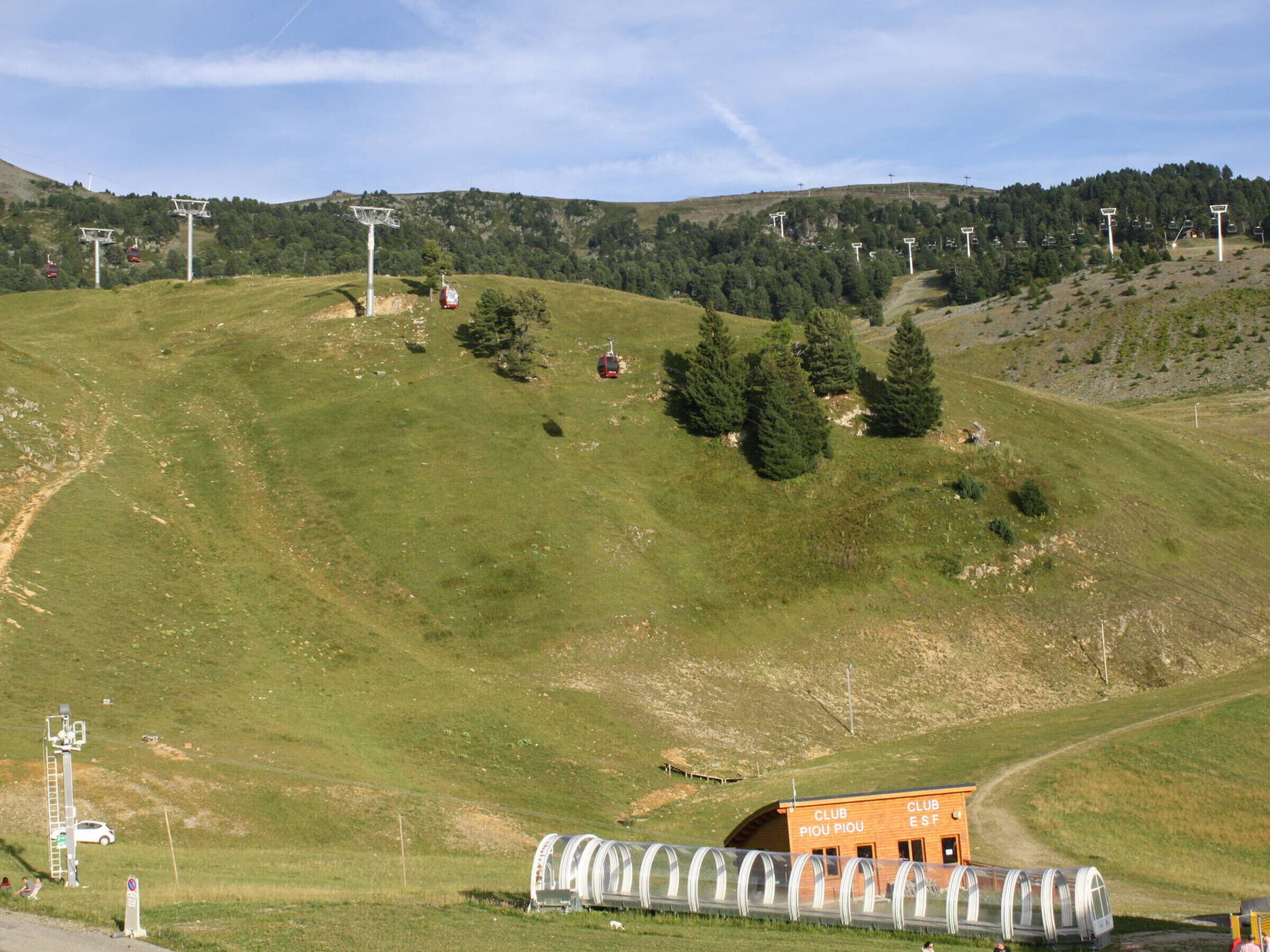 A hilly landscape with a ski lift, a wooden building labeled 'CLUB PIOU PIOU,' and a transparent structure, possibly a covered walkway or tunnel.