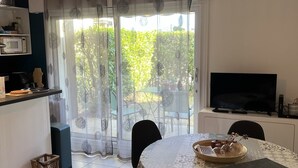 A dining area with a round table, chairs, a TV, and a kitchen area with a microwave and a shelf. - Central 1BR Garden Apartment, Close to Beach, Parking & Pet-Friendly (Cabourg)