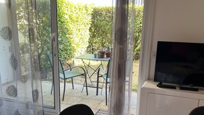 A room with a sliding glass door leading to a patio with a table and chairs, a television on a cabinet, and a patterned tablecloth on the dining table. - Central 1BR Garden Apartment, Close to Beach, Parking & Pet-Friendly (Cabourg)