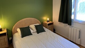 A bedroom with a green wall, a wicker headboard, a white bedspread, two bedside tables with lamps, and a window with curtains. - Central 1BR Garden Apartment, Close to Beach, Parking & Pet-Friendly (Cabourg)