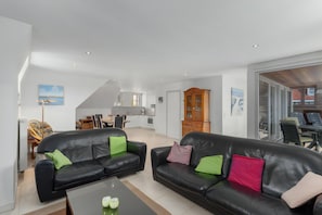 A modern living room with two black leather sofas, a glass coffee table, and a dining area in the background.