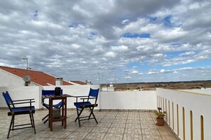 Een terras met een tafel, stoelen, een bloempot en een uitzicht op een verafgelegen landschap onder een bewolkte lucht.