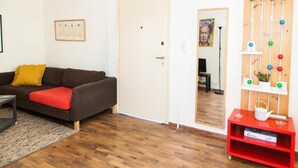 Un salon avec un canapé marron foncé, un coussin rouge, un plancher en bois, une porte blanche, une étagère rouge contenant des livres et un jeu de balles fixé au mur.