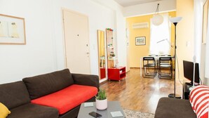 Un salon avec un canapé marron, un coussin rouge, une petite table sur laquelle se trouve une plante, et un téléviseur.