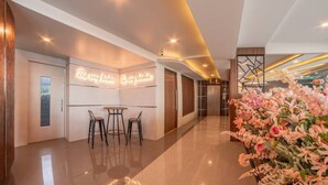 A modern hotel lobby with a reception desk, two bar stools, a large floral arrangement, and a polished floor. - The Ring Border (Sadao)