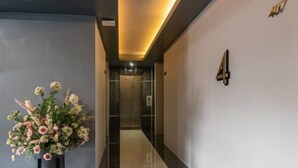 A hallway with a floral arrangement on the left, a number '407' on the right wall, and an elevator in the background. - The Ring Border (Sadao)