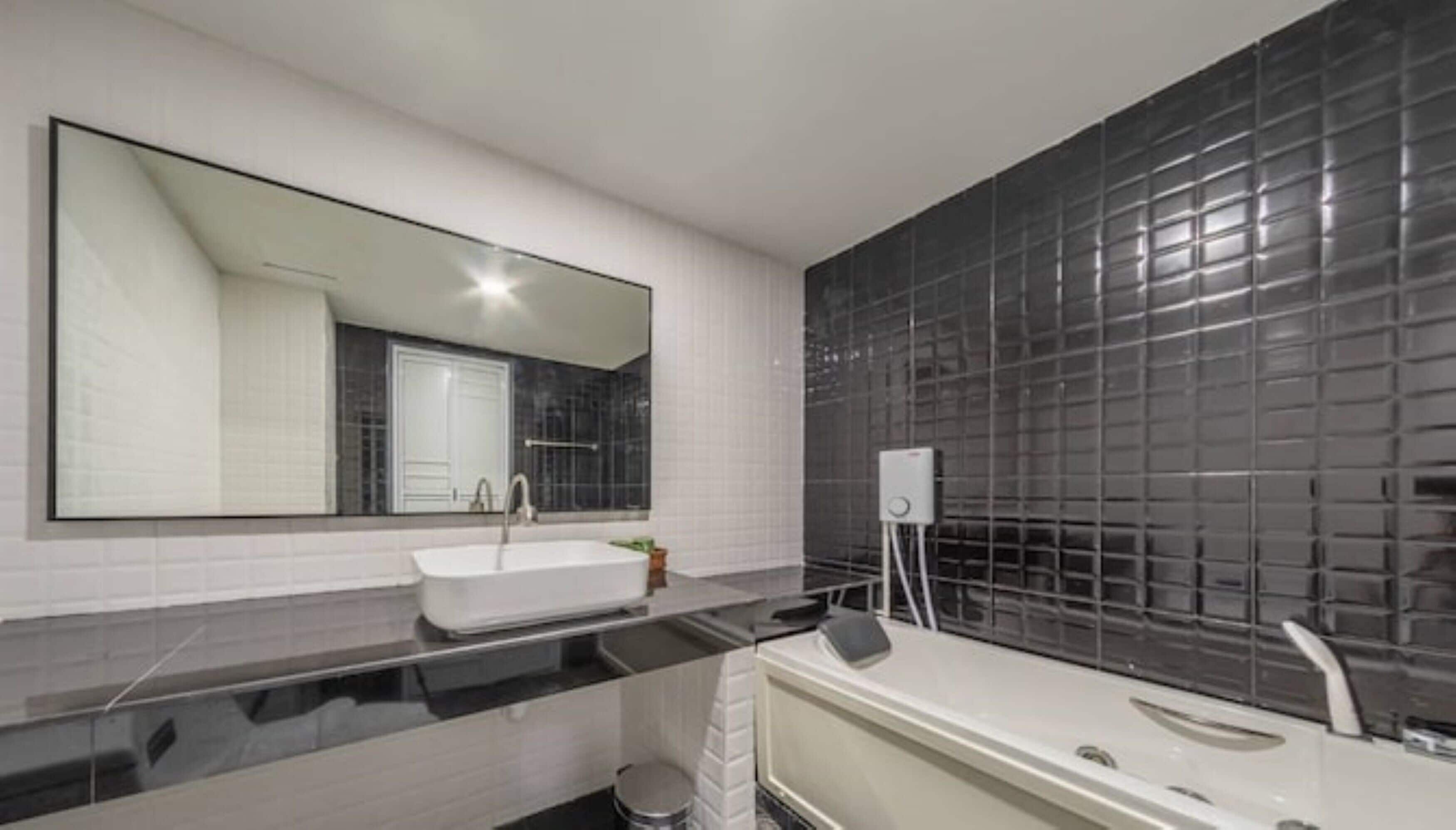 Un baño moderno con un espejo grande, un lavamanos blanco, una encimera negra y una bañera blanca, además de un revestimiento de pared de baldosas negras.