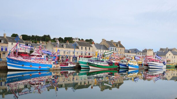 Un port où de nombreux bateaux sont amarrés, se reflétant dans l’eau calme, et entouré de bâtiments ornés de drapeaux et de banderoles.
