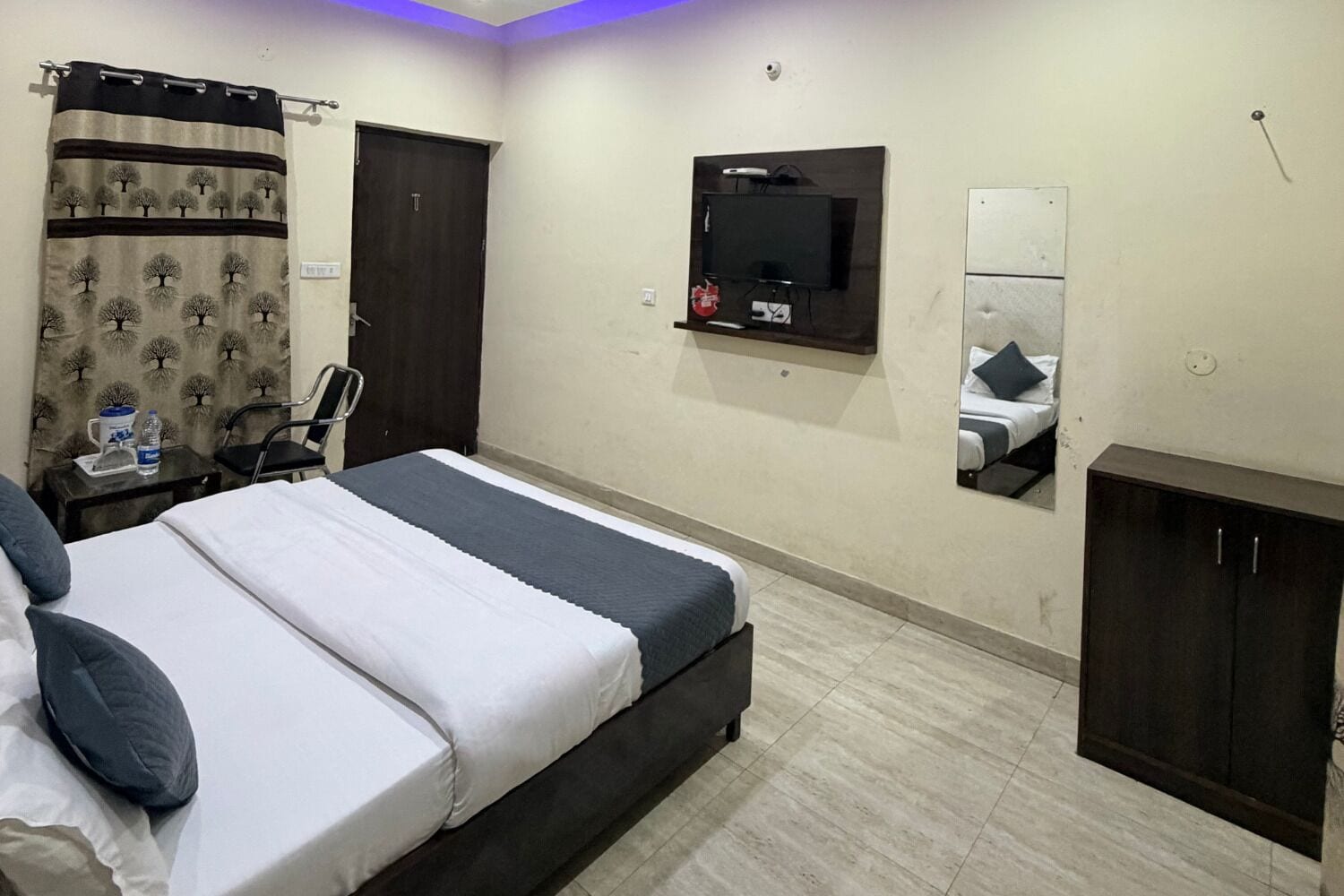 A hotel room with a bed, a TV mounted on the wall, a mirror, a wardrobe, and a curtain with a tree pattern.