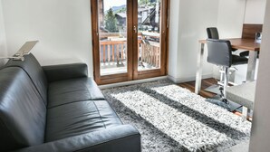 Uma sala de estar moderna com um sofá de couro preto, uma mesa de jantar de madeira com uma cadeira e uma grande porta de vidro deslizante que dá para uma varanda com vista para prédios e árvores.
