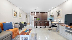 A modern living room with a sofa, coffee table, and a kitchen area with a dining table and bar stools. - Eden on the Bay 150 by HostAgents (Cape Town)