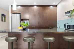 Cozinha moderna com armários de madeira, geladeira de aço inoxidável, espaço de bar com banquetas e uma planta suspensa.