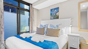A neatly made bed with blue and white bedding, a painting above it, and a small bedside table with a lamp. - Eden on the Bay 150 by HostAgents (Cape Town)