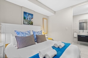 A modern hotel room with a large bed, a painting on the wall, a bathroom with a mirror and sink. - Eden on the Bay 150 by HostAgents (Cape Town)
