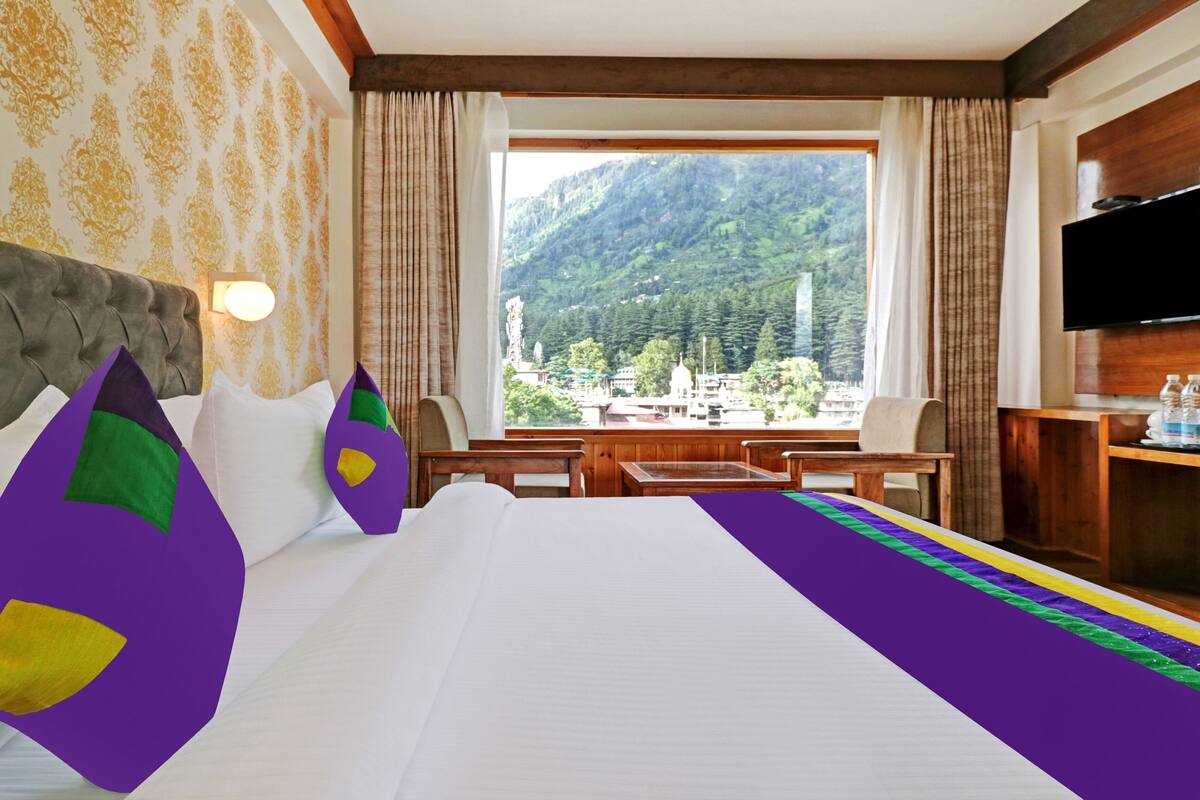 Habitación de hotel con una cama grande, cabecera de madera, vista a las montañas, un televisor de pantalla plana y una mesita de noche de madera con botellas de agua.